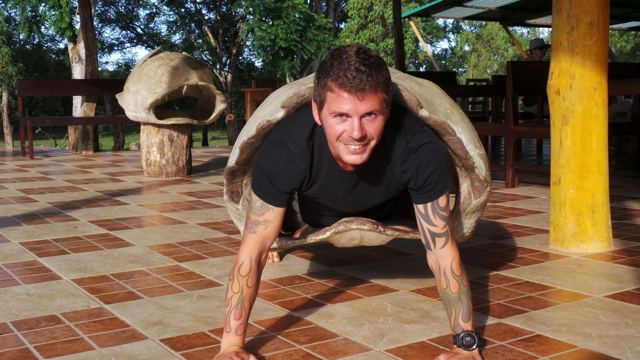 A man on all fours wearing an empty giant tortoise shell on his back.
