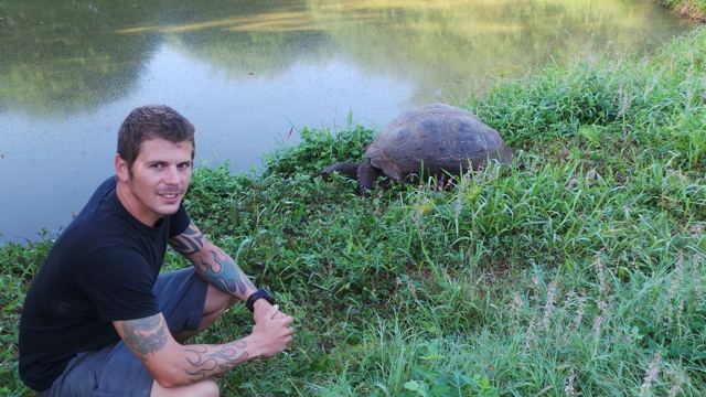 A man crouching on grass near a pond with a giant tortoise grazing beside him.