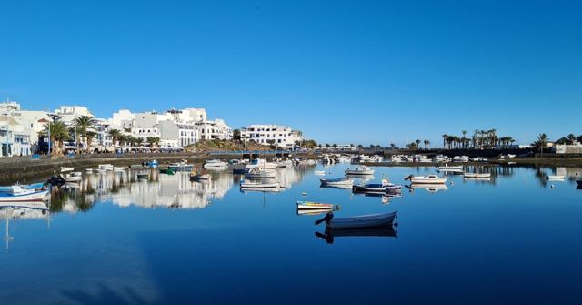 White buildings and small boats reflected in perfectly still blue water under a clear sky.