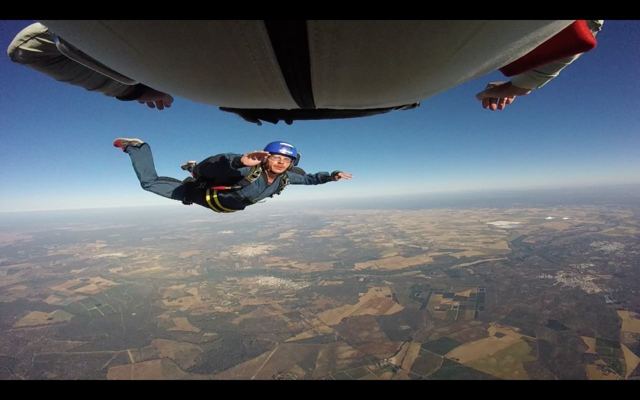 A skydiver in freefall below a plane wing with patchwork fields far below.