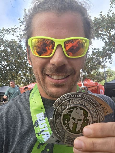 Smiling man in bright green sunglasses holding up a Spartan Race medal on a green lanyard.