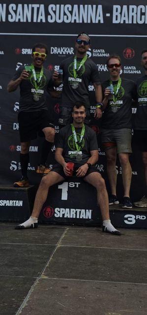 A man sitting on a first-place podium with other racers holding medals behind him.