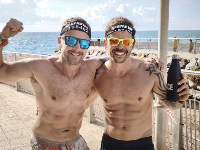 Two shirtless men in race headbands and sunglasses posing by the sea.
