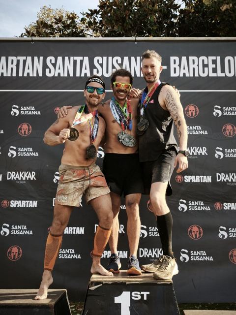 Three racers with medals posing on the podium in front of a Spartan backdrop.