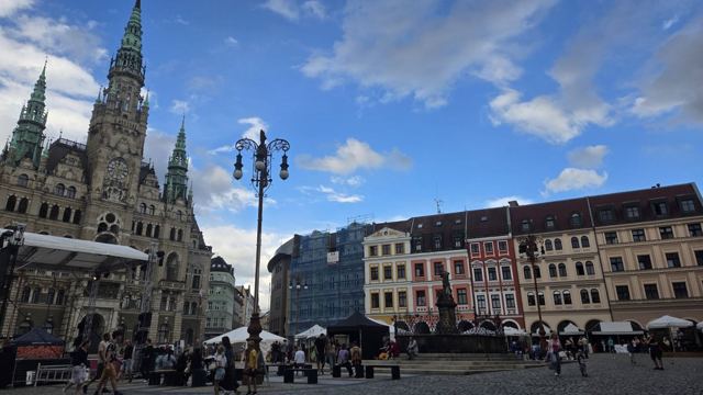 Ornate town hall with spires beside colourful buildings on a busy square under blue sky.