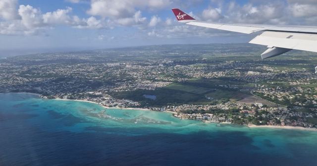 An aerial view of Barbados' coast with turquoise water, seen past a plane's wing tip.
