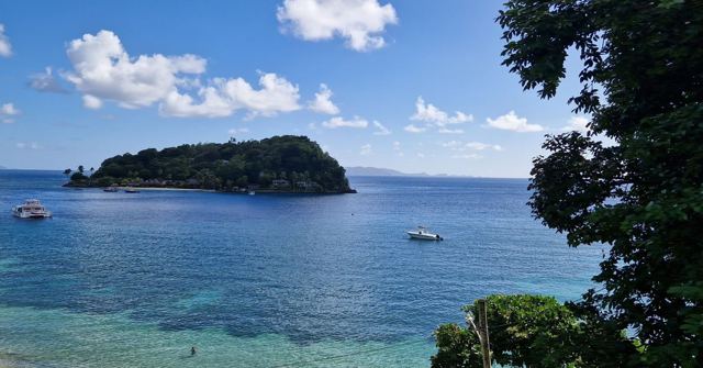 A small forested island in clear blue Caribbean water with boats anchored nearby.