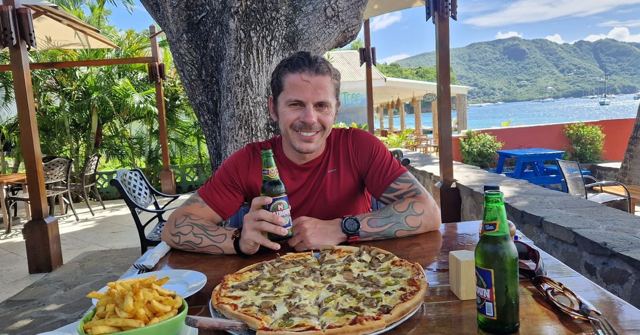 A smiling man at an outdoor table with a pizza, chips and beer, with a hillside harbour behind.