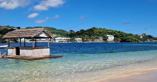 A thatched-roof hut on a platform in clear shallow water with green hills and houses behind.