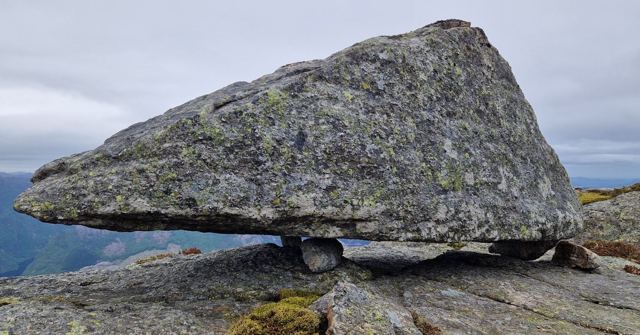 A huge angular boulder balanced on small rocks atop a mountain under a grey sky.