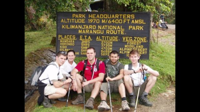 A group of hikers sitting in front of the Kilimanjaro National Park headquarters sign on the Marangu route.