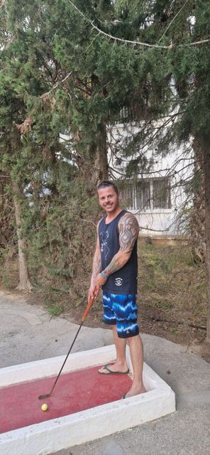A man in a vest and swim shorts standing in a garden under trees.