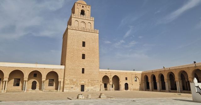 Tall square stone minaret rising above arcaded walls of a mosque courtyard.