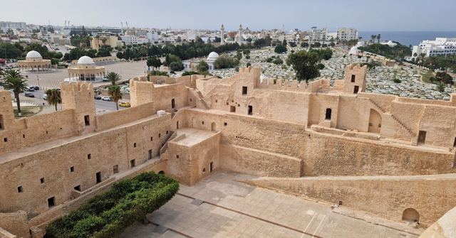 Sandstone fortress walls and courtyards with a white town and the sea in the distance.