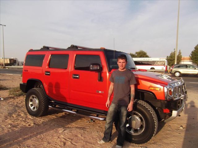 A man standing beside a red Hummer on sandy ground at sunset.