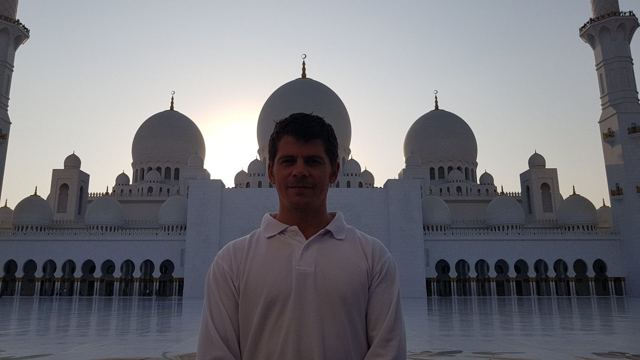 A man standing in front of the white domes and minarets of the Sheikh Zayed Grand Mosque at sunset.