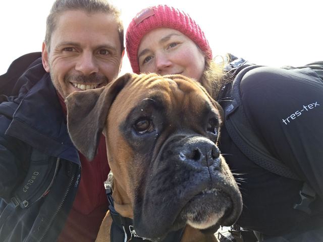 A man and a woman taking a selfie with a boxer dog in the foreground.