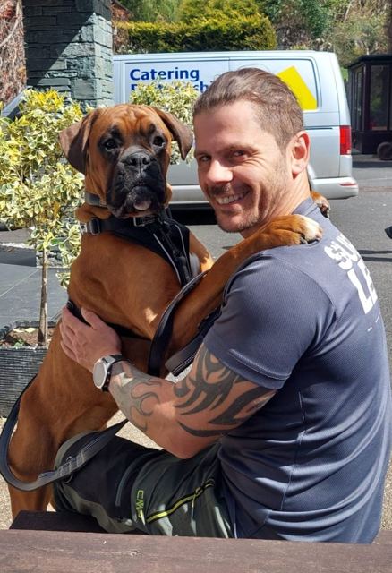 A smiling man hugging a big brown boxer dog outside.