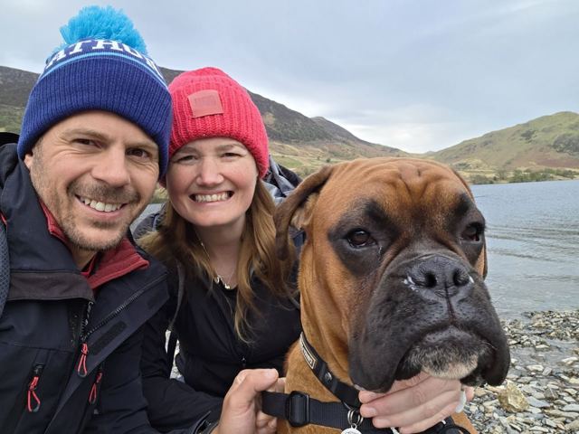 A man and a woman in beanies taking a selfie with a boxer dog by a lake.
