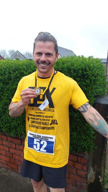 A smiling man in a yellow T-shirt holding up a medal, wearing a race number.