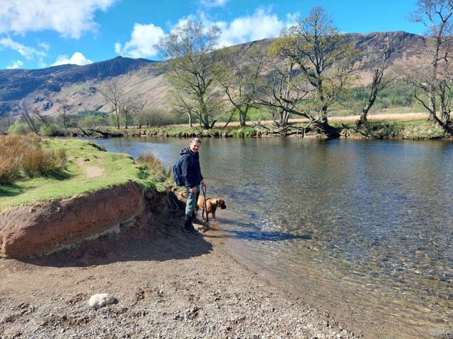 A person with a dog by a clear river with trees and hills behind on a sunny day.