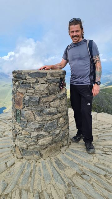 A man in a T-shirt standing beside the stone summit pillar of Snowdon.