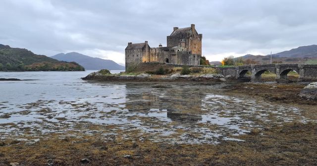 A stone castle on a small island linked by an arched bridge, with low tide revealing seaweed.
