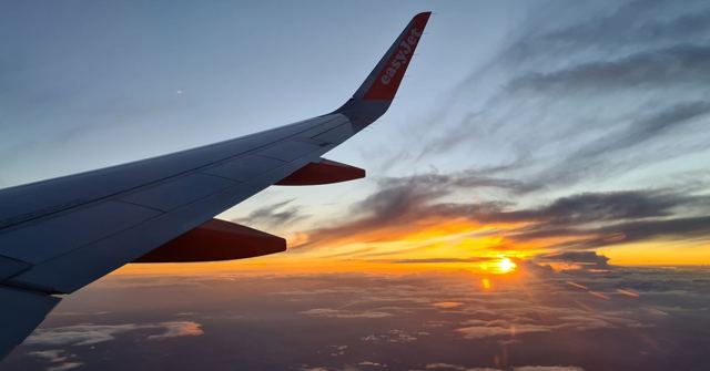 An aircraft wing above the clouds with a bright orange sunset on the horizon.