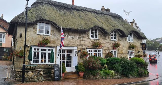 A thatched cottage pub with white windows, hanging flower baskets and a Union flag on a rainy street.
