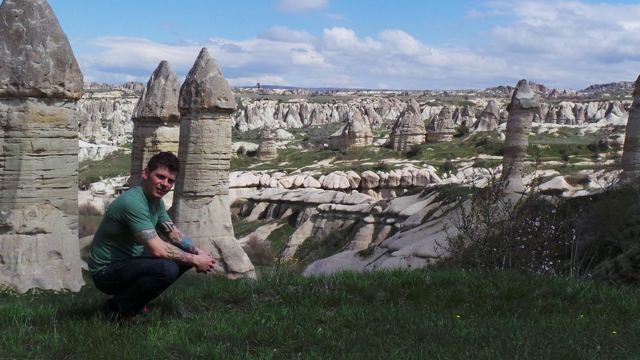 A man crouching on a grassy ridge above the rock chimneys and valleys of Cappadocia.