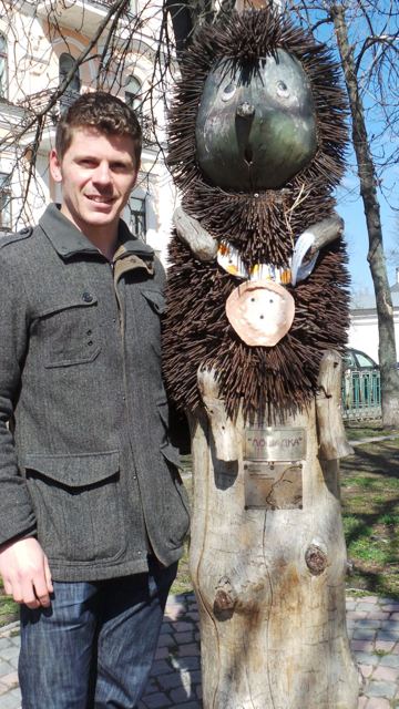 A man in a grey coat standing next to a whimsical wooden sculpture in a park in Kyiv.