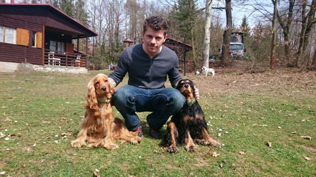 A man crouching on a lawn with two spaniels beside a wooden cabin.