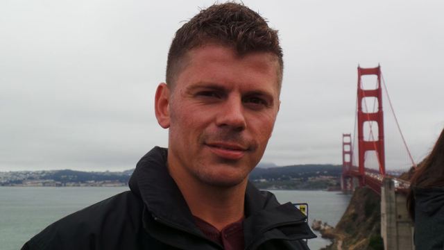 A man in a dark jacket in front of the Golden Gate Bridge on a hazy day.