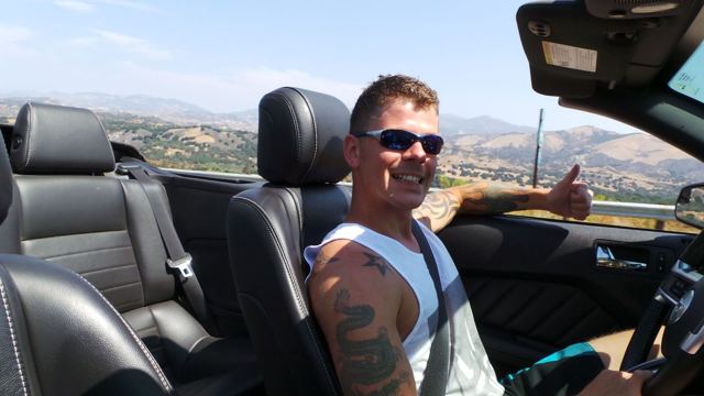 A man in sunglasses driving a convertible with dry hills behind.
