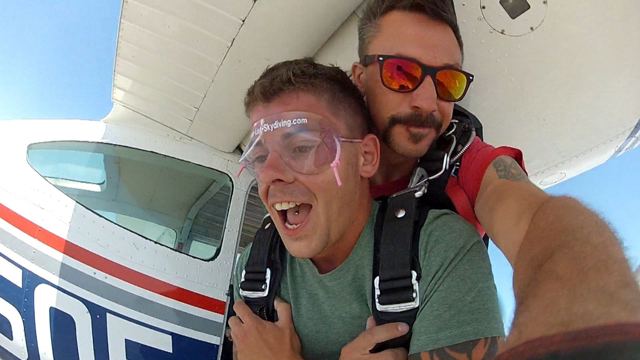 A man in goggles and a tandem harness grinning in the open door of a small plane, his instructor behind.