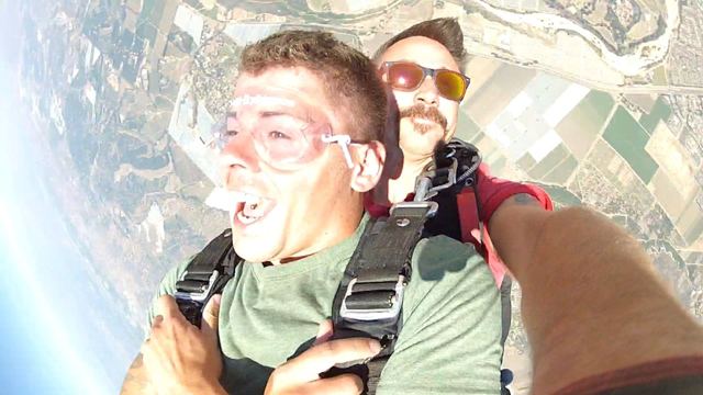 A man in goggles shouting in freefall during a tandem skydive over patchwork fields.