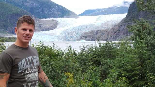 A man standing in front of the Mendenhall Glacier flowing down between forested mountains.