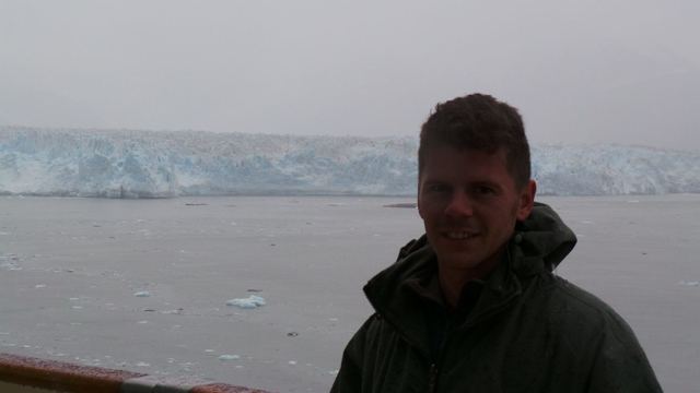 A man in a jacket on a ship's deck in front of the vast ice face of Hubbard Glacier.