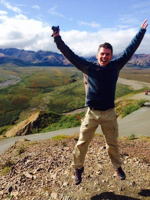 A man jumping with arms raised on a hilltop above a wide valley and mountains in Denali.