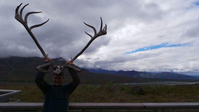 A man holding a pair of caribou antlers above his head against a cloudy sky.