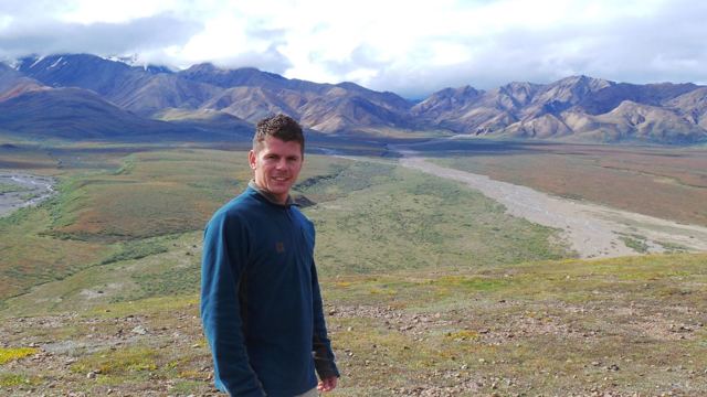 A man standing on a hillside above a wide river valley and mountains in Denali.
