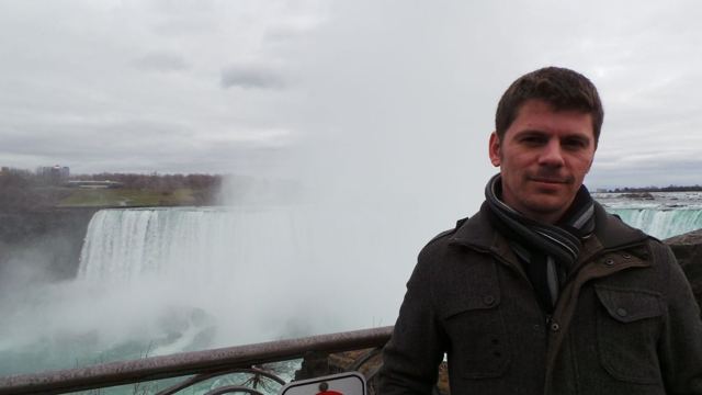 A man in a winter coat in front of the misty Horseshoe Falls at Niagara.