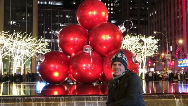 A man in a beanie posing beside a fountain full of giant red Christmas baubles in New York.