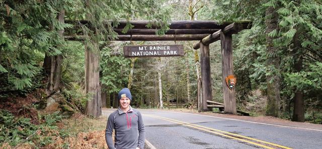 A man standing on a road beneath the wooden entrance arch of Mount Rainier National Park.