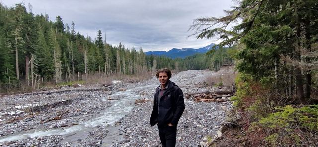 A man standing on a rocky riverbed lined with tall evergreen trees.