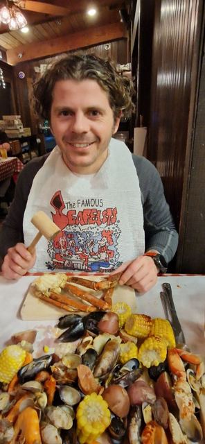 A smiling man wearing a paper bib holding a wooden mallet over a pile of crab legs.