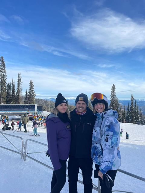 A man with two women in ski gear standing in front of ski slopes and pine trees.