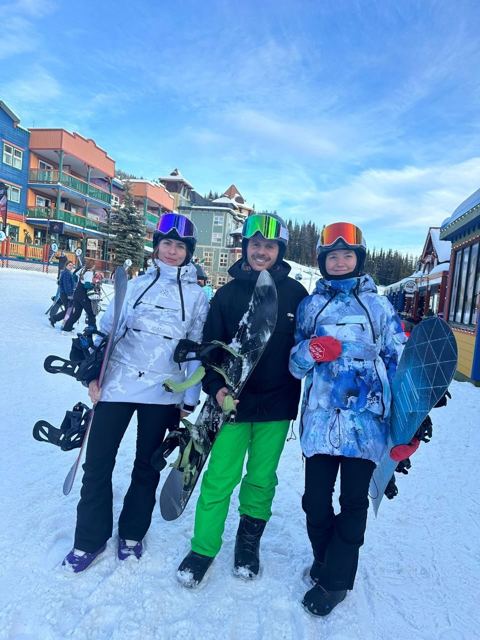 Three people holding snowboards in a snowy mountain village with colourful buildings.