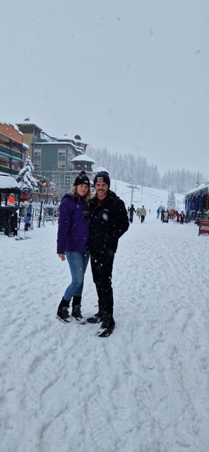 A man and a woman in winter coats standing in falling snow in a ski village.