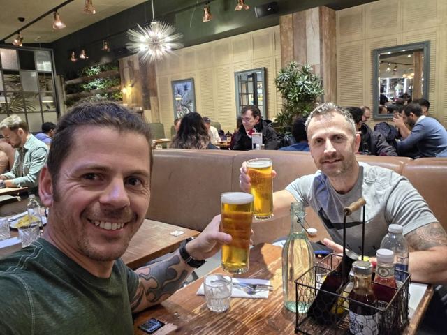Two men toasting with pints in a busy airport restaurant.